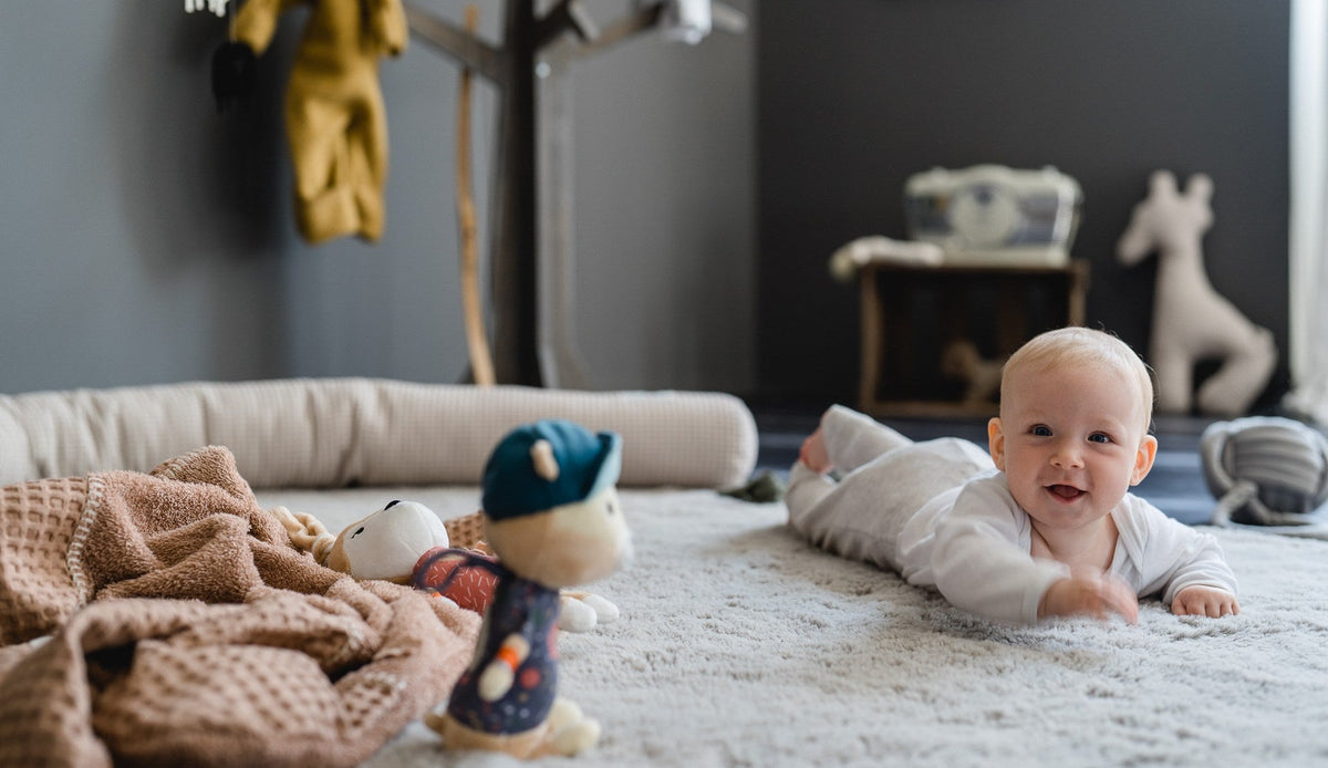 Lächelndes Baby liegt auf dem Bauch auf einem weichen Teppich im Kinderzimmer und streckt die Hand nach einem Kuscheltier aus