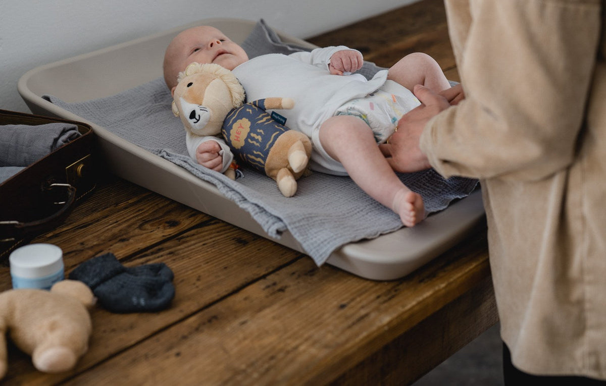 Wach liegendes Baby auf dem Wickeltisch, daneben der Löwe Eddie, ein Elternteil hält sanft das Bein