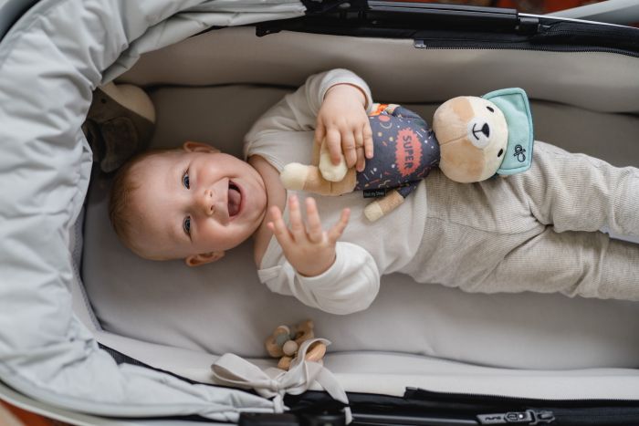 Baby liegt lachend im Kinderwagen und hält Jay der Bär